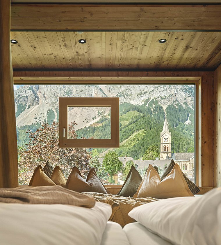 Panorama-Ausblick vom Bett auf Kirche und Berge durch großes Fenster mit Holzrahmen
