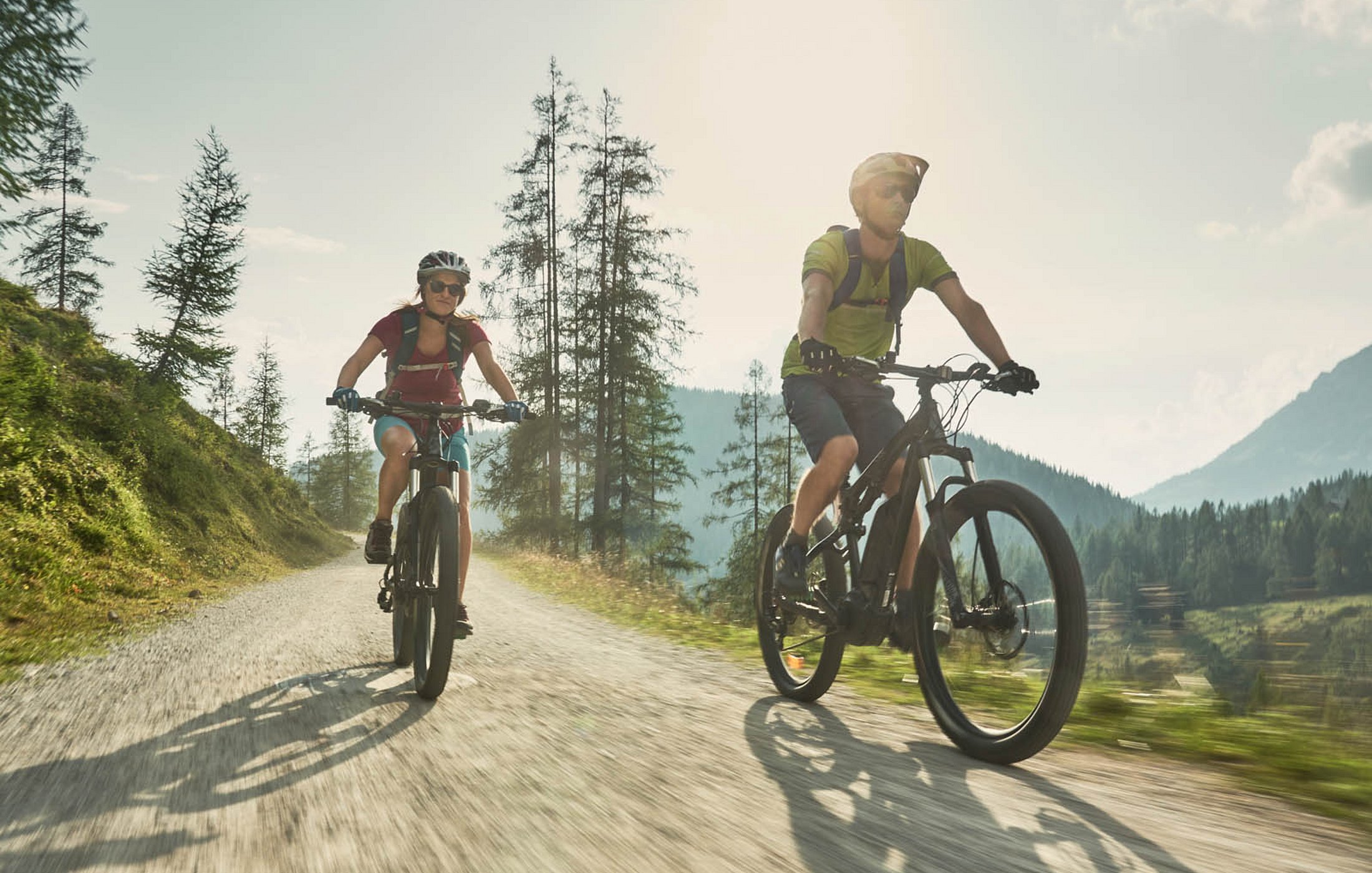 Paar mit Helm fährt auf E-Bikes durch sonnigen Waldweg in den Bergen