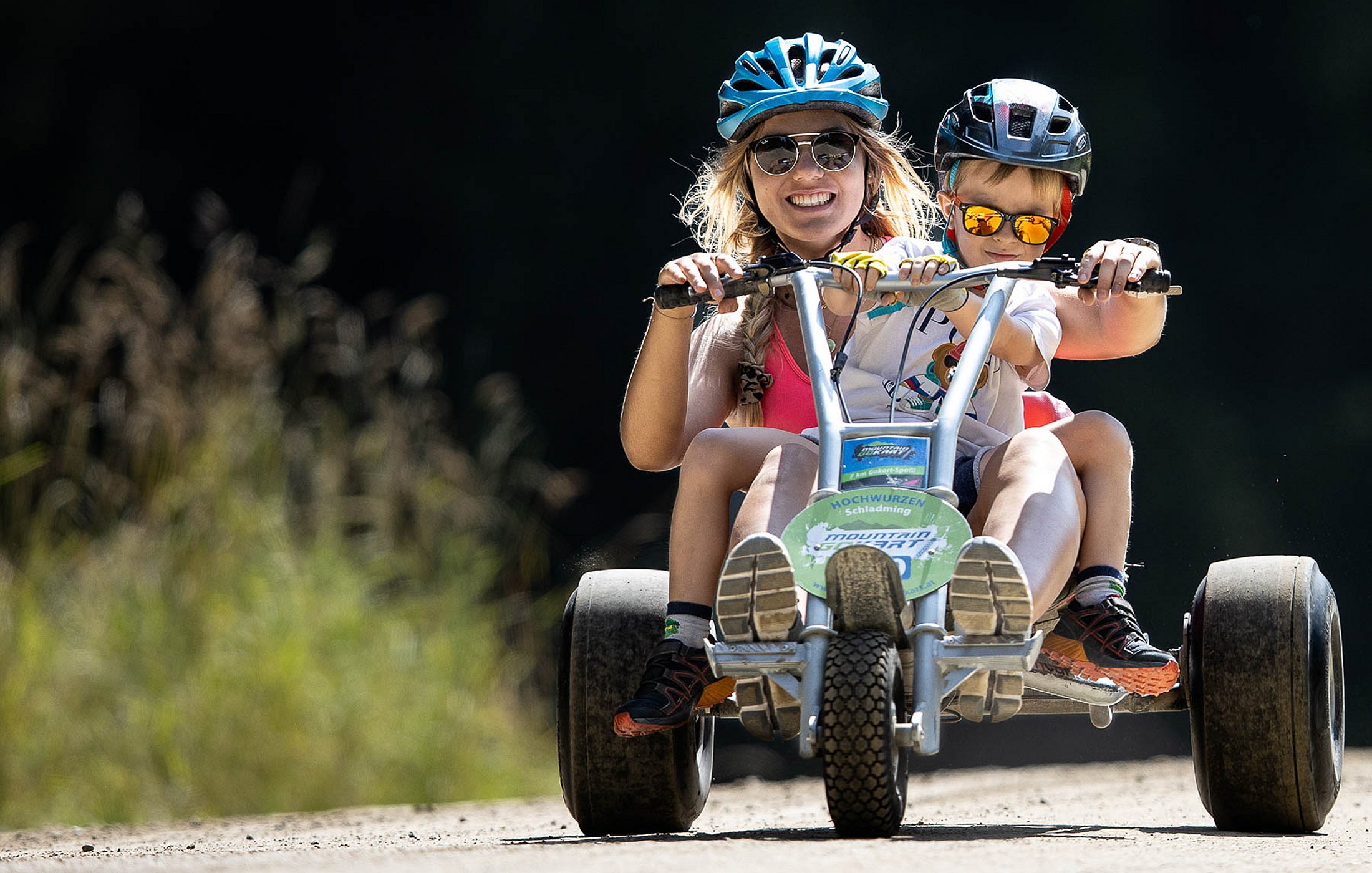 Eine lachende Frau mit blauem Helm und Sonnenbrille fährt zusammen mit einem kleinen Jungen, der ebenfalls einen Helm und Sonnenbrille trägt, auf einem Mountain-Gokart einen Schotterweg hinunter