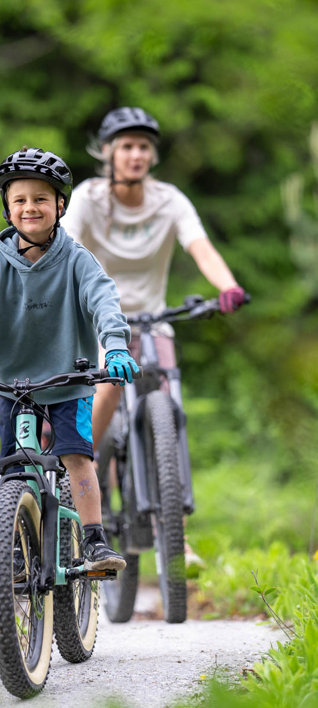 Nahaufnahme eines lächelnden Kindes auf einem mintgrünen Mountainbike auf einem sonnigen Pfad, im Hintergrund eine Frau auf einem Fahrrad