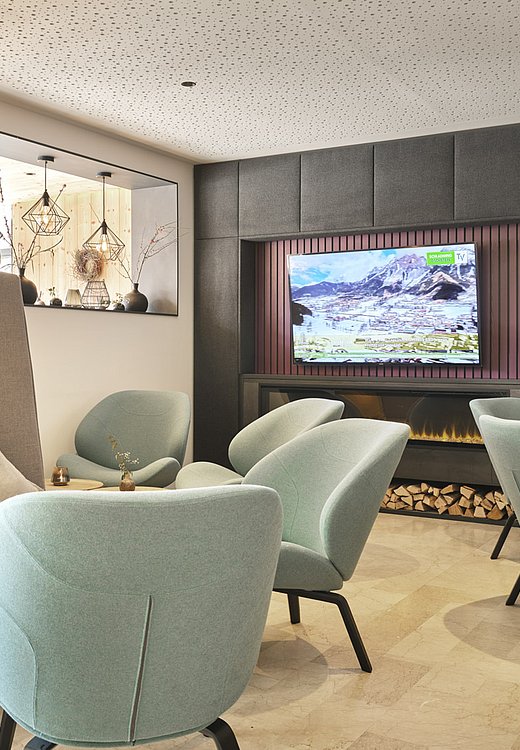 Loungebereich mit Fernseher, Kamin und hellblauen Sesseln vor grauer Wandverkleidung