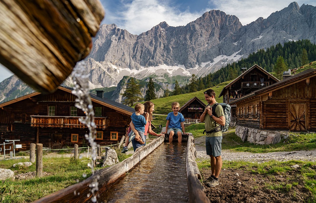 Eine vierköpfige Familie rastet an einem langen hölzernen Wassertrog bei urigen Almhütten