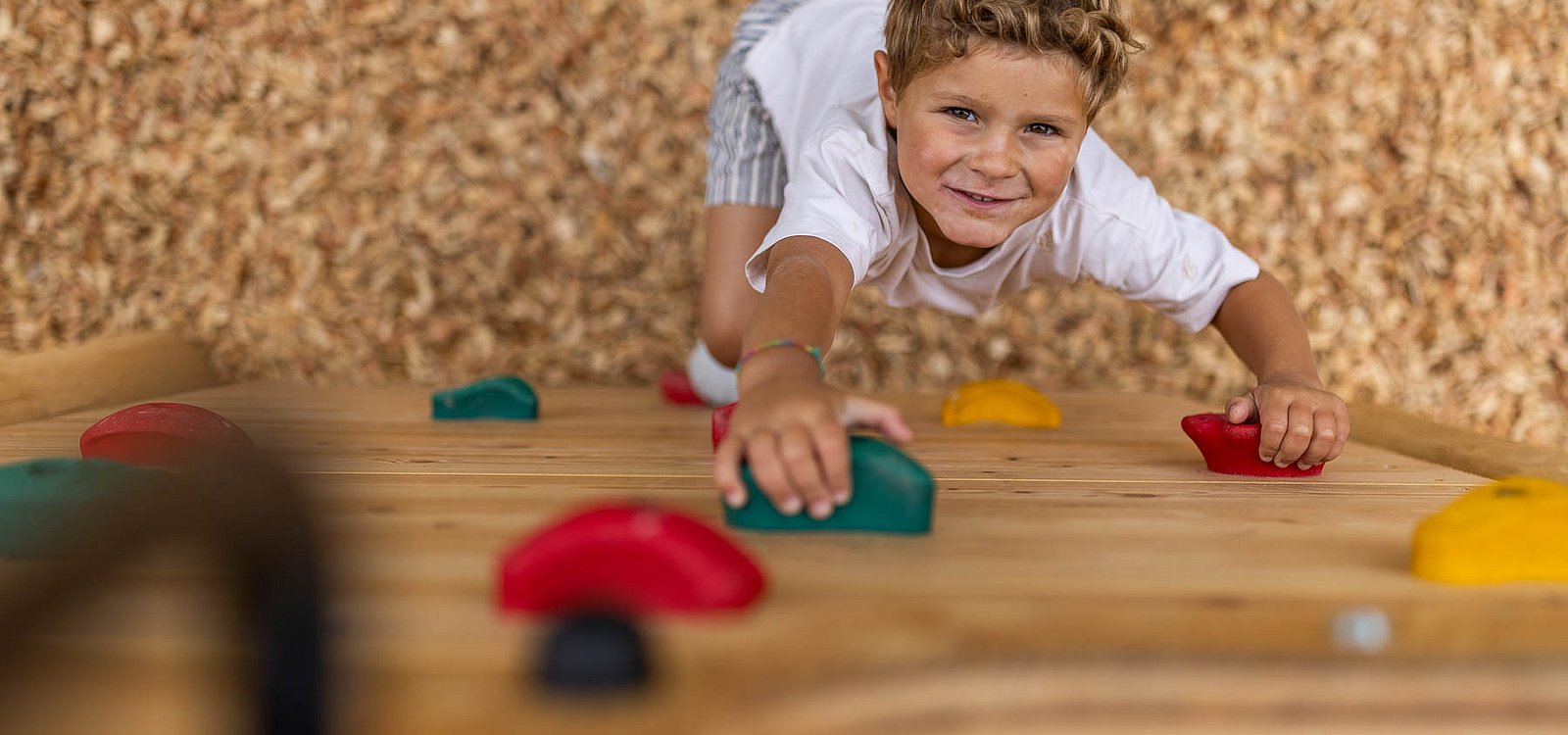 Ein kleiner Junge greift beim Klettern an einer hölzernen Kletterwand nach den bunten Haltegriffen