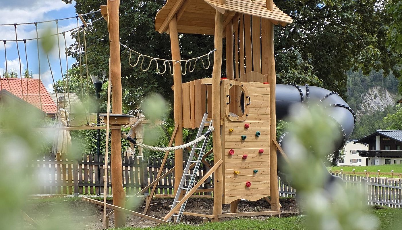 Hölzerner Spielturm mit grauer Röhrenrutsche und Kletterwand, umrahmt von grünen Blättern