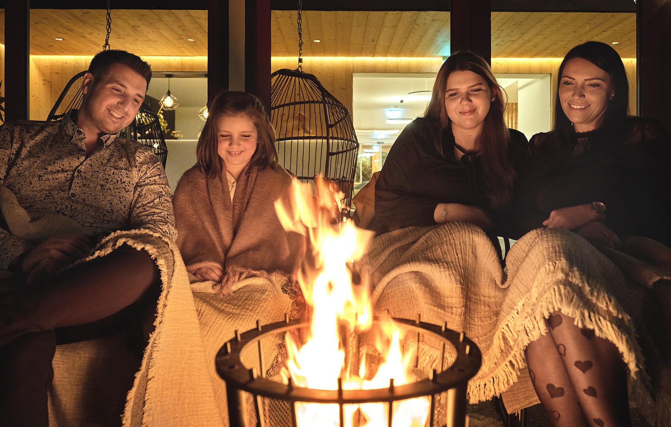 Eine vierköpfige Familie sitzt am Abend in warme Decken gehüllt glücklich um eine brennende Feuerschale im Freien