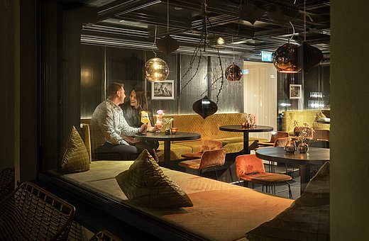 Couple sitting in the cosy hotel bar in the evening, enjoying colourful cocktails beneath atmospheric pendant lights