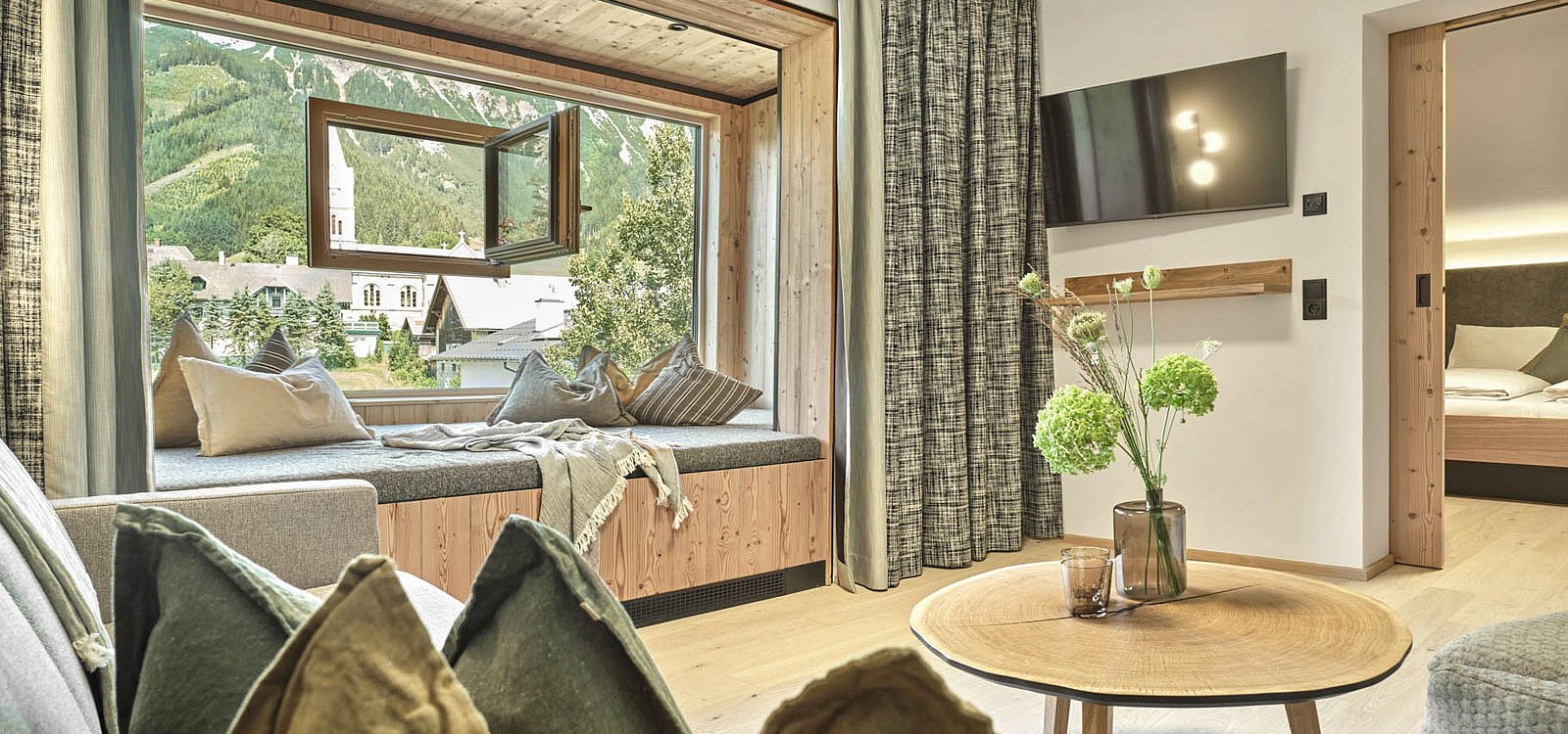 Wohnbereich mit großem Fenster, Sitzbank und Blick auf Berge und Kirchturm