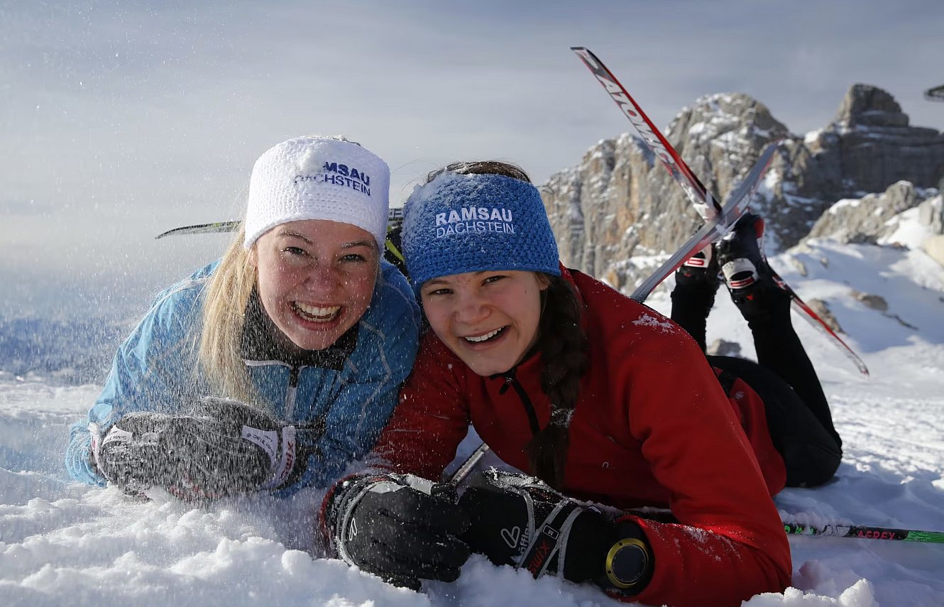 Zwei lachende Mädchen liegen im Schnee mit Skiern vor winterlicher Bergkulisse