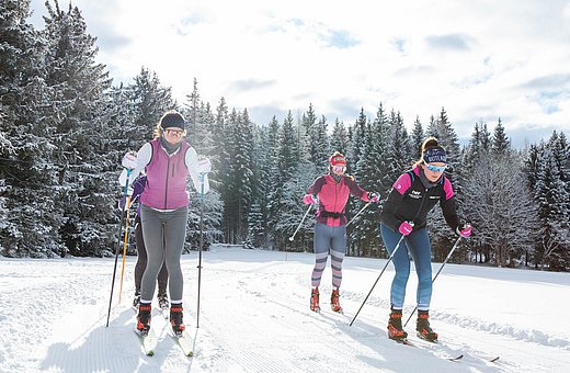 Drei Frauen beim Langlaufen auf einer präparierten Loipe im verschneiten Wald