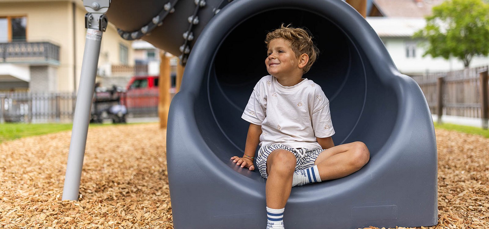 Ein kleiner Junge sitzt lächelnd am Ende einer dunklen Röhrenrutsche auf einem Spielplatz