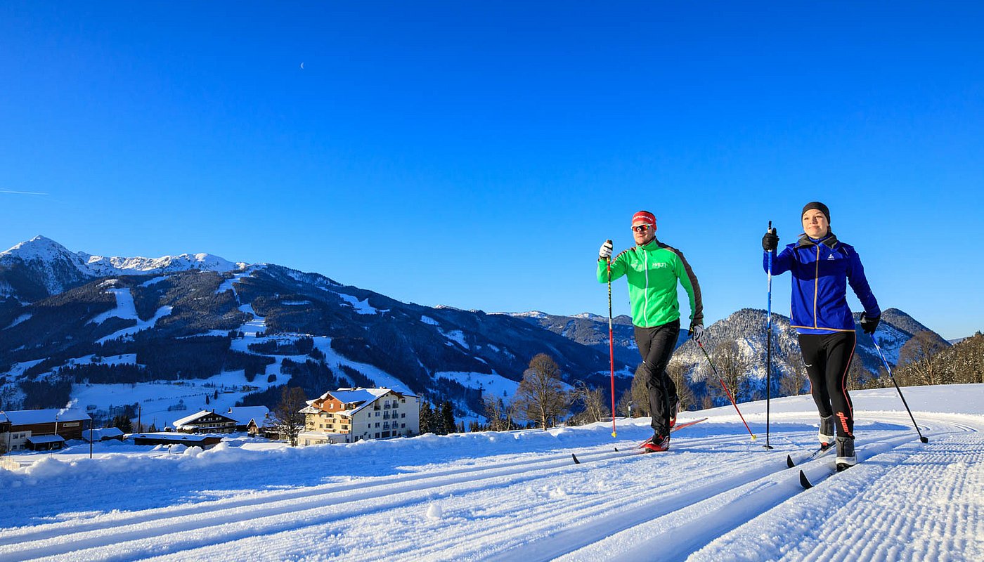 Zwei Langläufer auf sonniger Loipe mit Blick auf verschneite Berge und tiefblauen Himmel