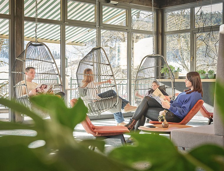 Familie liest Zeitschriften in hängenden Korbsesseln und einem Loungesessel im lichtdurchfluteten Wintergarten mit Schneeblick