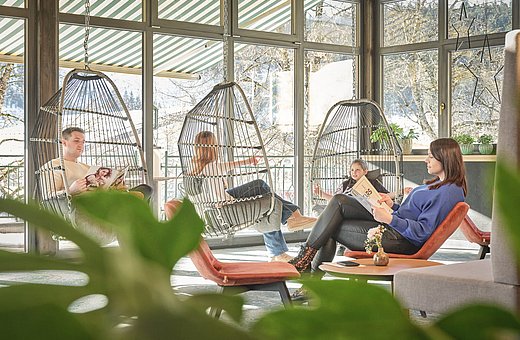 Familie liest Zeitschriften in hängenden Korbsesseln und einem Loungesessel im lichtdurchfluteten Wintergarten mit Schneeblick