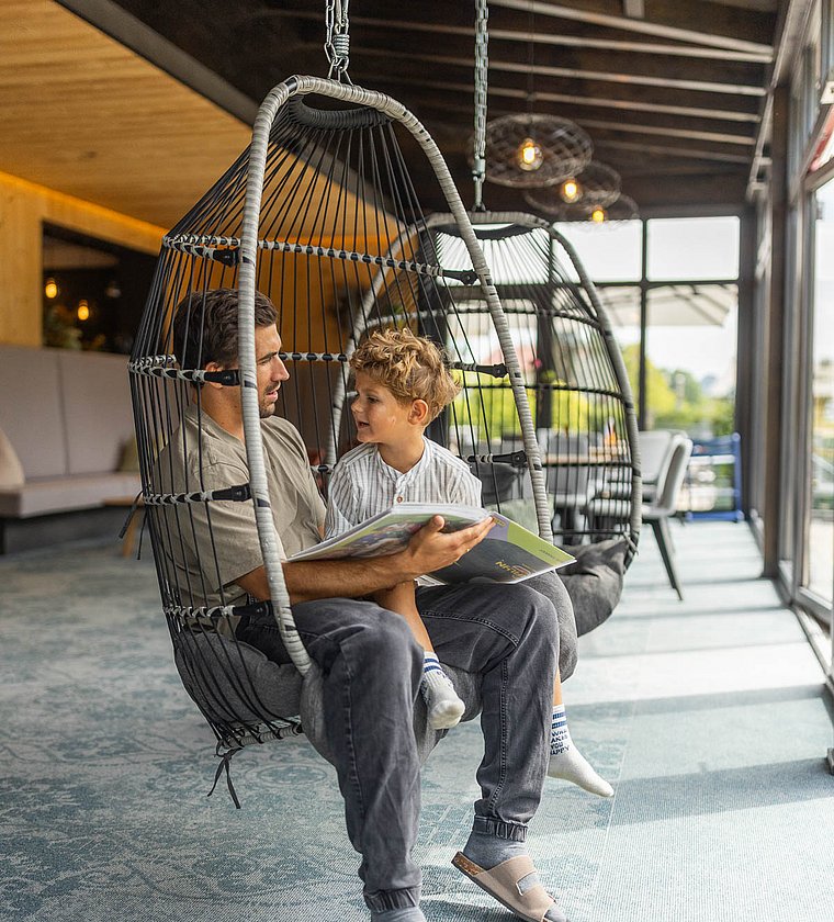 A father sitting with his son in a hanging chair reading him a book