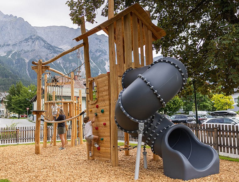 A young boy climbing the wooden wall of a play tower with a tube slide in front of mountains