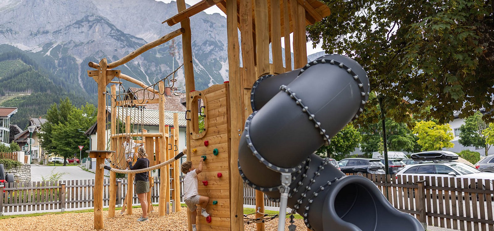 Ein kleiner Junge klettert an der Holzwand eines Spielturms mit Röhrenrutsche vor Bergkulisse