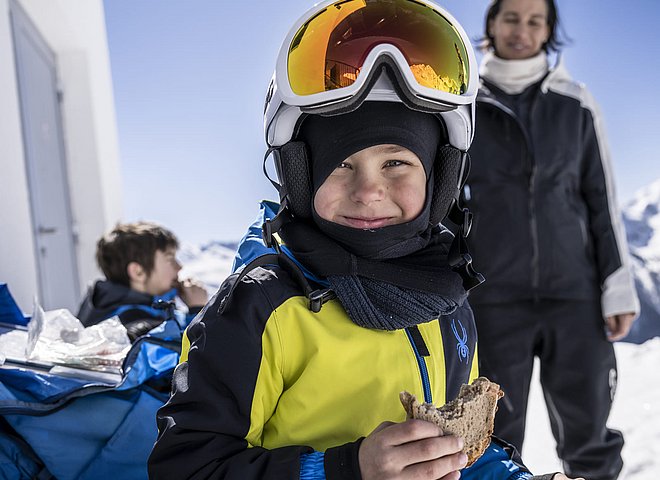 Junge im gelben Skianzug mit Helm und Brille isst Brot vor schneebedeckten Bergen beim Skitag