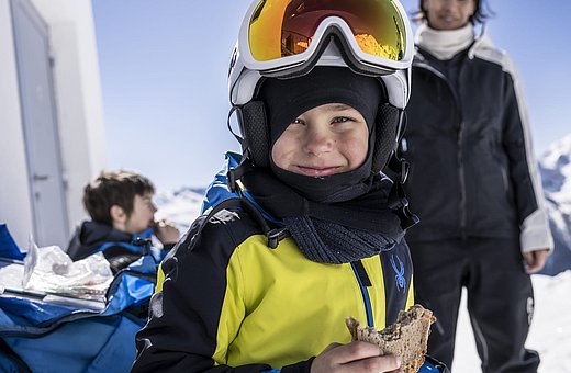 Junge im gelben Skianzug mit Helm und Brille isst Brot vor schneebedeckten Bergen beim Skitag