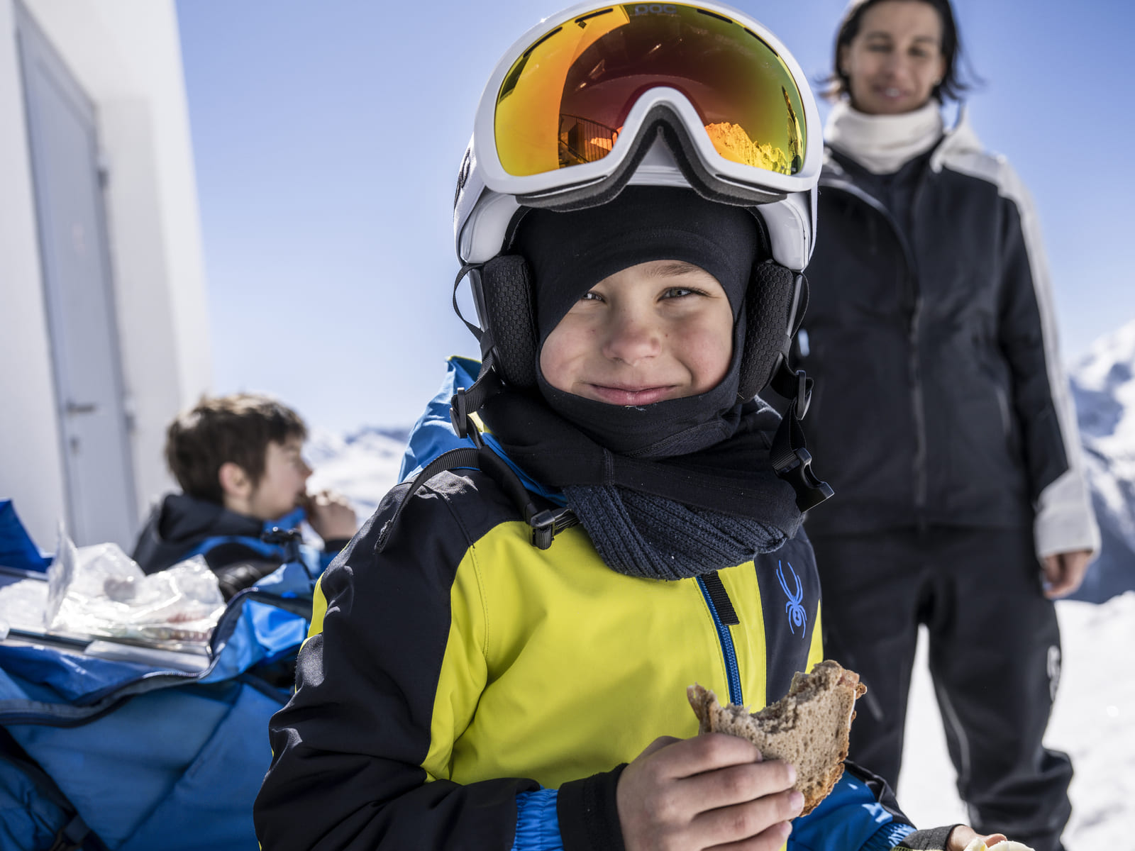 Junge im gelben Skianzug mit Helm und Brille isst Brot vor schneebedeckten Bergen beim Skitag